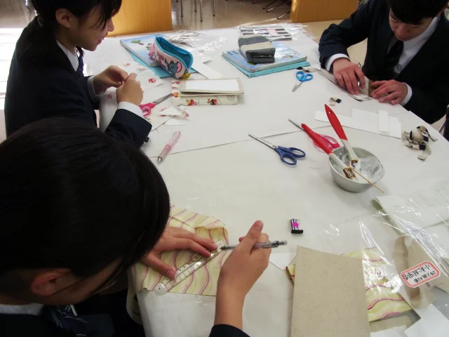 家庭科教材の導入事例：中学2年生の家庭科授業にてカルトナージュのノートカバーを約4時間かけて作成する生徒たちの様子：布を型紙に合わせて切る生徒たちの写真