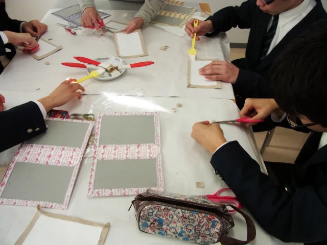 家庭科教材の導入事例：中学2年生の家庭科授業にてカルトナージュのノートカバーを約4時間かけて作成する生徒たちの様子：素材をノリで貼り付ける様子の生徒達の写真